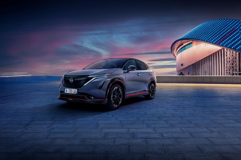 Vue de face 3/4 d'une Nissan Ariya NISMO bleu-gris avec des accents de design rouge sur un ciel bleu-gris et avec un bâtiment en arrière-plan