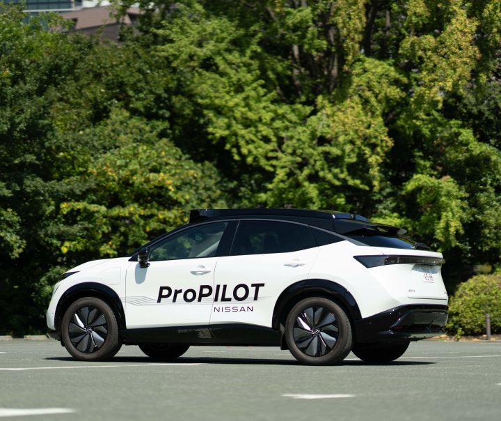 3/4 Heckansicht eines weißen Nissan Ariya mit der schwarzen Aufschrift ProPILOT Nissan, der vor dicht begrünten Bäumen steht