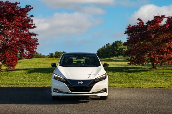 Front view of a white 2025 Nissan LEAF parked in front of bright green grass, blue skies, and vibrant red trees