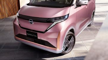 建物のある屋外スペースで、ピンク色の「日産サクラ」を右前方から斜めに捉えたクローズアップ画像。フロントフェイスから側面にかけてが強調され、背景はややぼかされて建築物の壁面が見える。