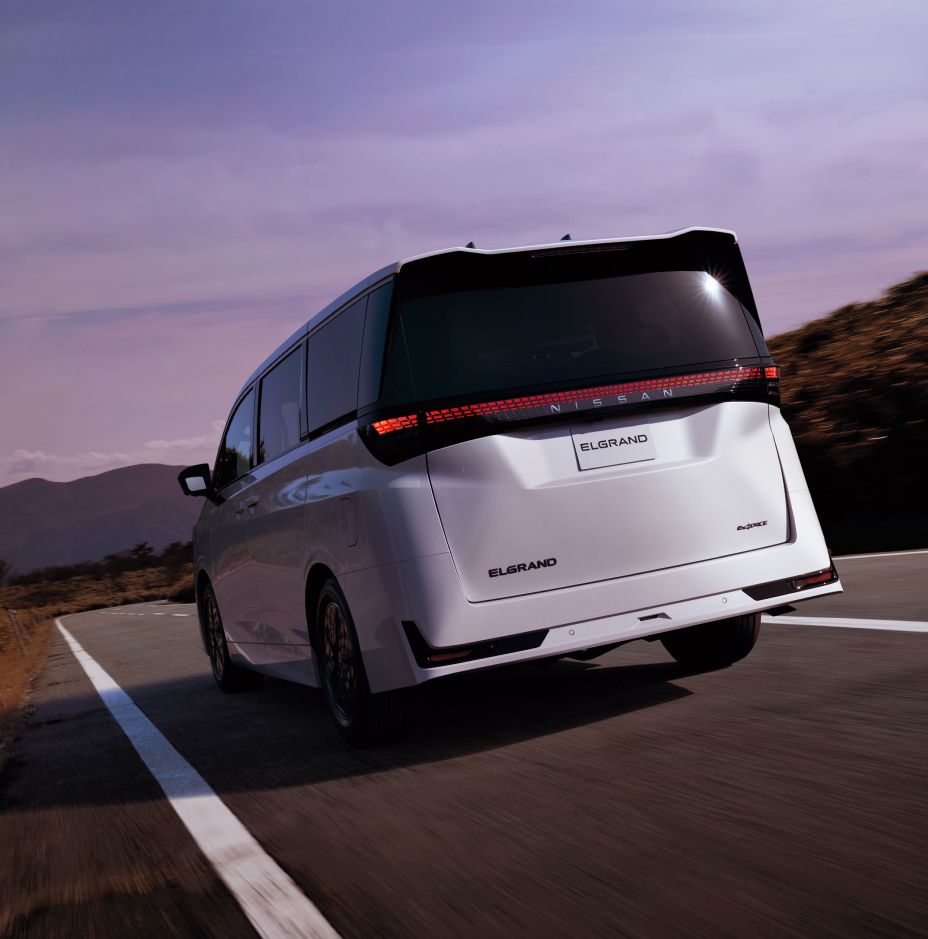 A white All new ELGRAND driving along a winding mountain road, viewed from the rear-left angle. The vehicle is traveling toward the right-front direction, featuring a black roof, dark-tinted windows, and a full-width red rear light bar that spans the tailgate. Mountains and low vegetation form the background beneath a soft purple sky, creating an open scenic setting that highlights the premium rear styling and bold presence of the minivan.