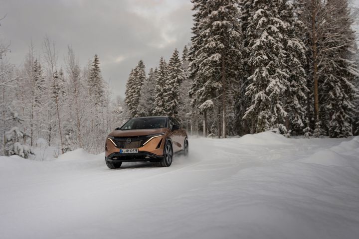 Elektroauto Nissan Ariya e‑4ORCE in Akatsuki Copper mit Dach und Außenspiegel in Pearl Black fährt auf schneebedeckter Straße mit verschneiten Bäumen auf beiden Seiten, 3/4 Front