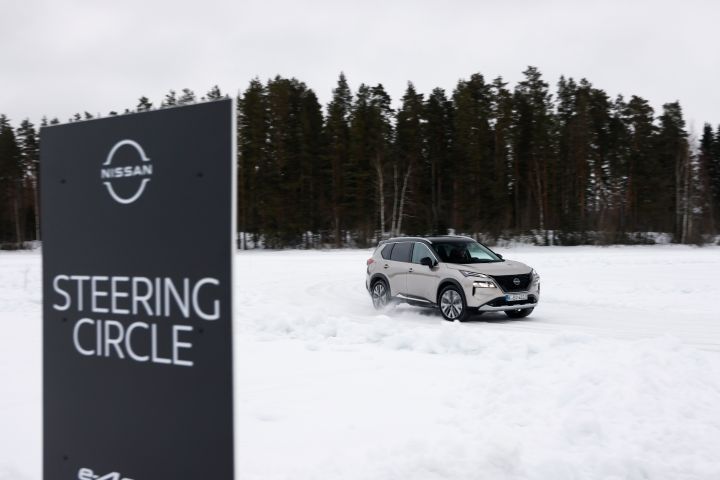Schwarzes Schild mit weißer Schrift Nissan e‑4ORCE Steering Circle steht im Vordergrund, im Hintergrund fährt ein silberner Nissan X‑Trail e‑Power e‑4ORCE auf Schnee