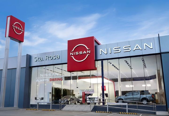Exterior of a Nissan dealership labeled “Nissan Sta. Rosa,” featuring a modern glass façade with large “NISSAN” signage and the red Nissan logo prominently displayed. Several vehicles are visible inside the brightly lit showroom, with an accessible ramp and steps leading to the entrance.