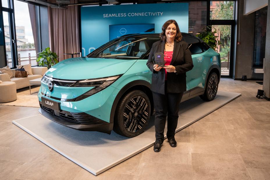 Katherine Zachary, VP Communications, steht vor einem türkisen Nissan LEAF, sie hält lächelnd ihren Women Automotive Network Award in den Händen
