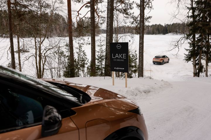 Im Vordergrund Teil eines Elektroautos Nissan Ariya e‑4ORCE in Akatsuki Copper mit Dach und Außenspiegel in Pearl Black und ein zweites im Hintergrund fahren auf einen schneebedeckten Eissee