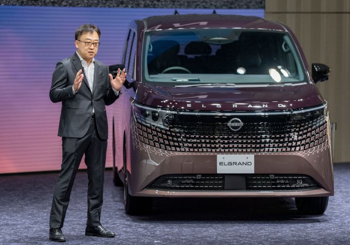 発表会場のステージで説明を行う執行職 杉本全と、新型エルグランド。画面左側にはダークグレーのスーツを着た執行職 杉本全が立ち、正面やや右方向を向きながら両手を上げて説明している。画面右側にはブラウン系の新型エルグランドが正面を向いて展示され、ワイドなフロントグリルと横一文字のヘッドランプが強調されている。背景にはピンクからブルーへと変化するグラデーションの大型LEDスクリーンが広がり、会場照明が車体表面に反射している。ステージ上で人物と車両を並べて紹介する、新型エルグランドの発表シーンを捉えた写真。