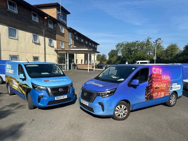 A pair of Nissan Primastar vans parked in front of a Travelodge hotel. The vans feature colourful Travelodge branding.