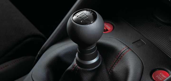 Close-up detail of the six-speed manual transmission shifter in a Fairlady Z NISMO. The black spherical shift knob features an engraved shift pattern for gears one through six and reverse on the top surface. The shifter is wrapped in a black leather boot with red contrast stitching, surrounded by a black center console. A red “START ENGINE STOP” button is visible beside the shifter, reinforcing the performance-oriented character of the cockpit. The image highlights the driver-focused controls and sporty interior detailing unique to the Fairlady Z NISMO.