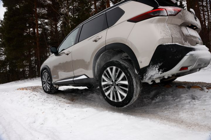 Heckansicht eines silbernen Nissan X‑Trail e‑Power e‑4ORCE, der über schneebedeckte Baumstämme fährt, ein Rad schwebt frei in der Luft
