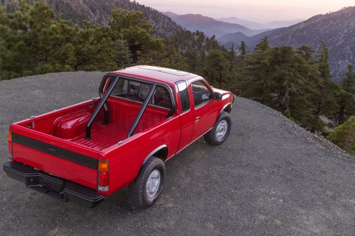 Angled high level view of a 1990 Nissan Hardbody 4x4 parked on a cliff overlooking pine trees.
