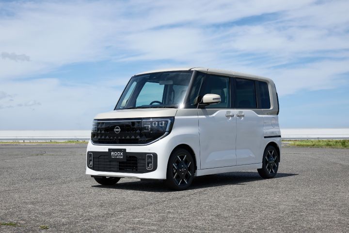The new Nissan Roox kei car, in white with a modern design featuring black front grille and wheels, is parked on an open asphalt surface. The background includes a clear blue sky with clouds, suggesting a coastal setting.