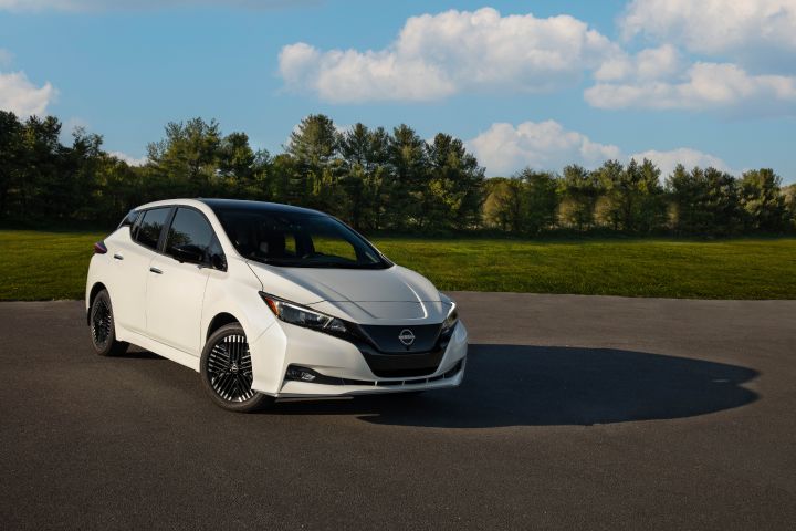 Front right 3/4 view of a white 2025 Nissan LEAF on a plain with blue skies and lush trees behind.