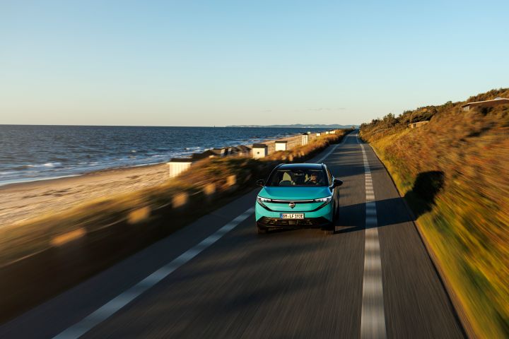Ein türkiser Nissan LEAF fährt frontal auf den Betrachter zu auf einer schmalen Straße, zu deren linken Seite sich Strand und Meer befinden