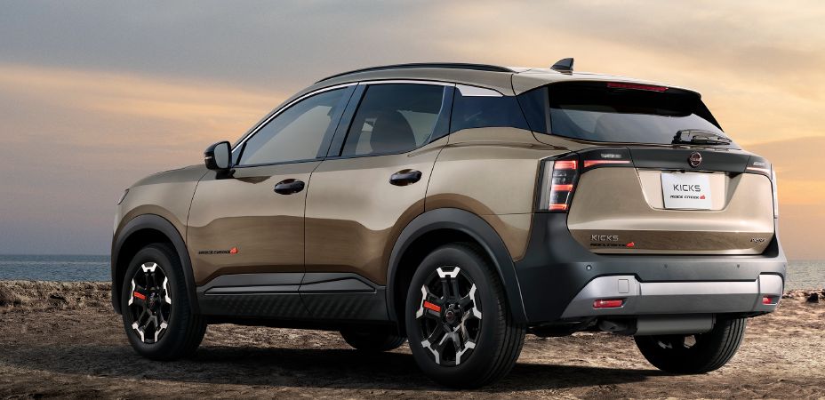 A beige-toned kicks rock creek is parked in a rear three-quarter view facing left. The black rear gate garnish, unique alloy wheels, and lava red accent parts reinforce its outdoor-inspired character. Distinctive vertical led rear combination lamps and muscular fender arches enhance its commanding suv presence. In the background, a calm ocean extends toward the horizon. The vehicle is positioned on rugged, rocky terrain, emphasizing the tough and active image of the kicks rock creek.