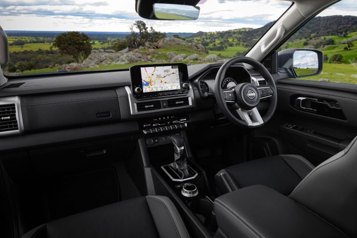 Wide front-cabin view of a 2026 Nissan Navara ST-X interior, showing the dashboard, steering wheel, centre console, automatic gear selector, and 9-inch Touch infotainment screen displaying a navigation map, with a rural landscape visible through the windscreen.