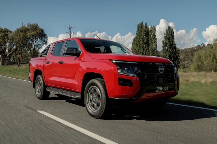 Front three‑quarter view of a 2026 Nissan Navara ST‑X dual cab painted in Outback Red, travelling along a sealed road with grassy hills and scattered trees in the distance.