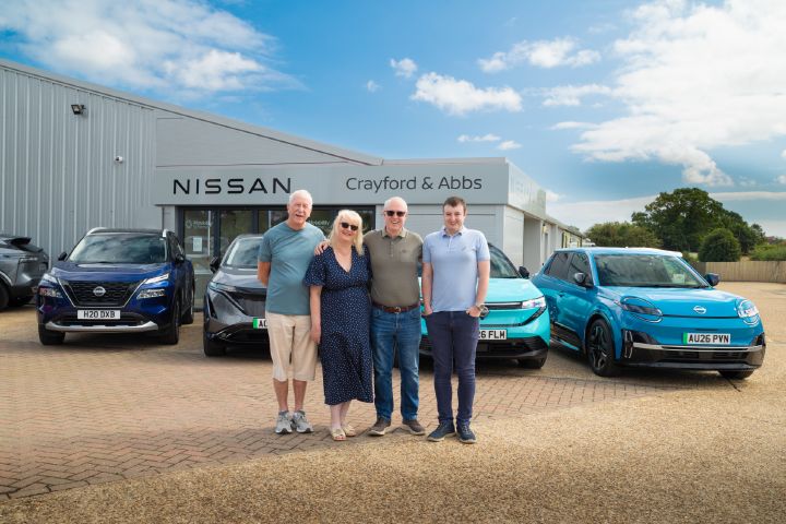 Group standing outside a Nissan dealership with a Nissan X‑Trail, Ariya, LEAF and MICRA parked behind them