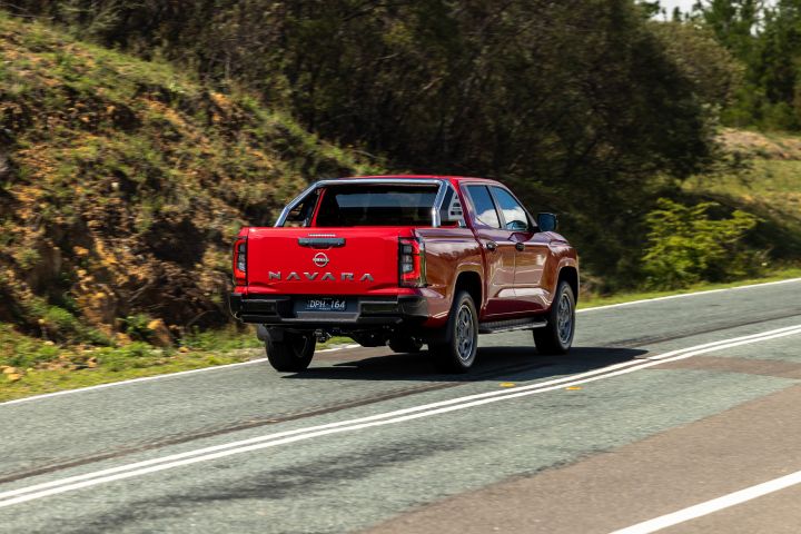Rear three‑quarter view of a red 2026 Nissan Navara ST‑X travelling on a rural highway next to a sloping embankment.