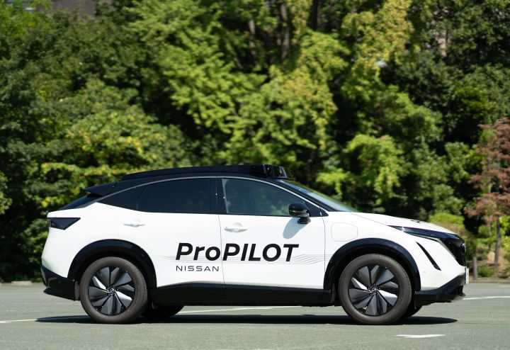Seitenansicht eines weißen Nissan Ariya mit der schwarzen Aufschrift "ProPILOT NISSAN", der auf einem Parkplatz vor grünen Bäumen steht
