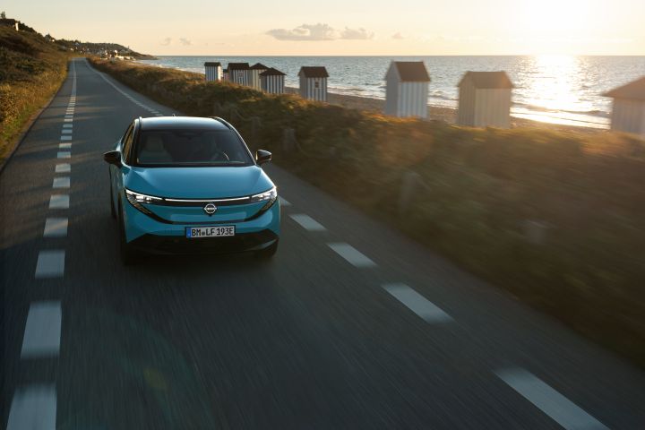Photo de face d'une Nissan LEAF turquoise roulant au coucher du soleil sur une route étroite longeant la plage.