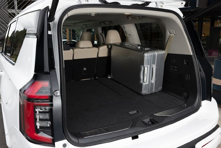 2026 Nissan Y63 Patrol finished in white, rear cargo area shown with the tailgate open, third-row seats folded flat and a large silver suitcase loaded inside.