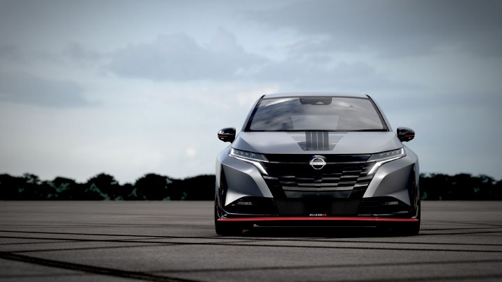 A silver and black two-tone sports car, the Aura Nismo RS Concept, parked on an outdoor skid pad in a full front view. The vehicle features a low stance and a sharp front design, with the Nissan logo on the center of the grille and a red accent line with the 'NISMO' logo on the lower bumper. The hood has a black stripe, and the background shows a cloudy sky and distant trees, with tire marks visible on the asphalt surface