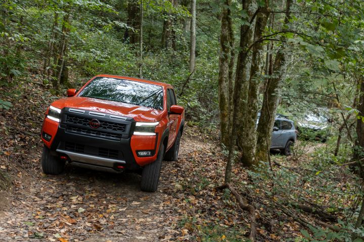 Two Nissan cars and a truck traverse through a winding path in leaf covered woods.