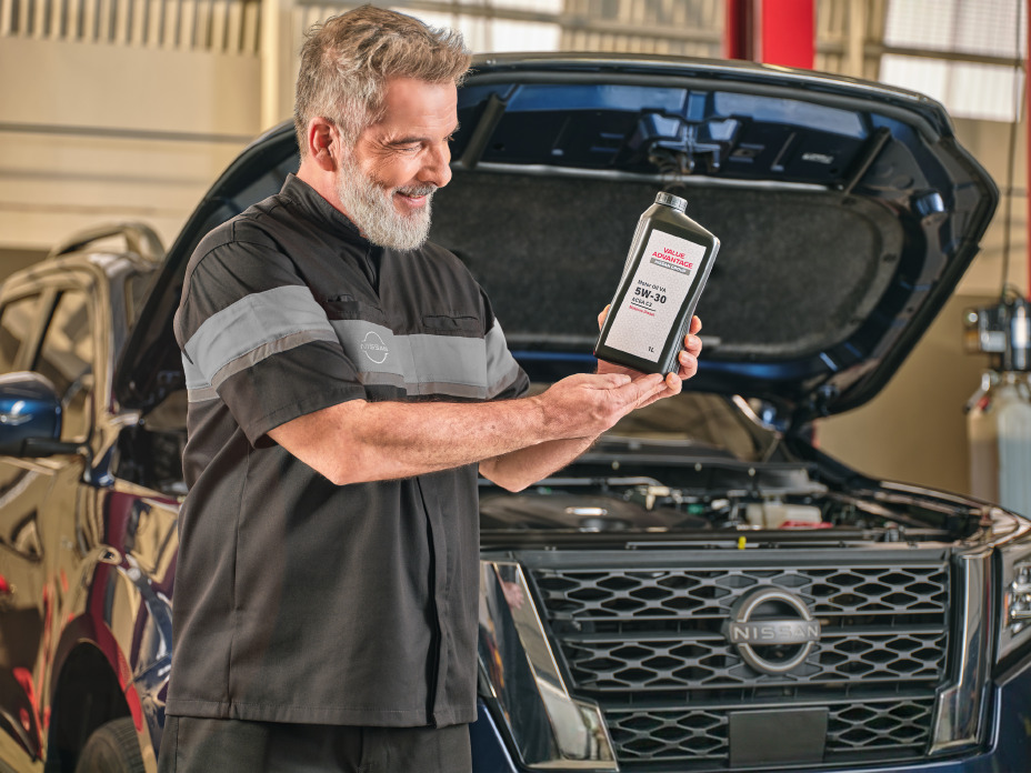 Reparador segura um frasco de óleo da Value Advantage à frente de uma Nissan Frontier azul com o capô aberto