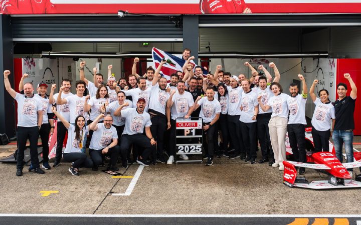 Grupo de pessoas com camisetas brancas comemorando em frente à garagem; ao centro, placa com os dizeres "OLIVER P1 2025", bandeira erguida e parte de um carro de corrida visível à direita