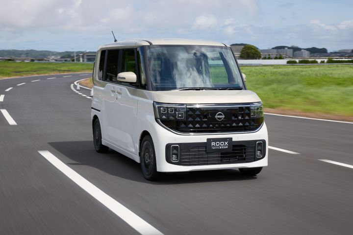 The new Nissan Roox kei car, featuring a white pearl / frozen vanilla pearl two-tone, is driving on a curved road. The background features green fields and buildings in the distance under a partly cloudy sky. The vehicle is moving forward, showcasing its outdoor usage and design.
