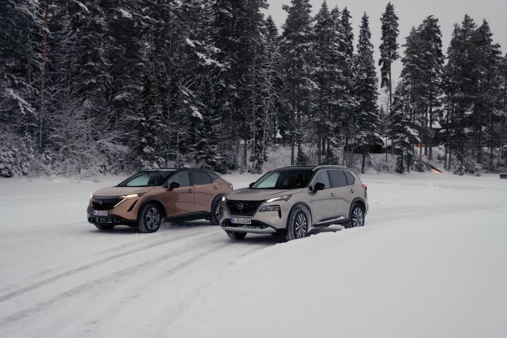 Silberner Nissan X‑Trail e‑Power e‑4ORCE und Elektroauto Nissan Ariya e‑4ORCE in Akatsuki Copper mit Dach und Außenspiegel in Pearl Black fahren nebeneinander auf schneebedeckter Straße mit verschneitem Wald im Hintergrund, 3/4 Front