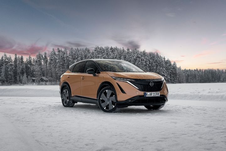 Elektroauto Nissan Ariya e‑4ORCE in Akatsuki Copper mit Dach und Außenspiegel in Pearl Black auf Eissee mit schneebedeckten Bäumen im Hintergrund, 3/4 Front