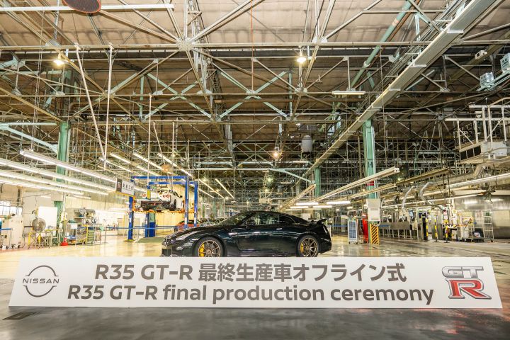 日産自動車 栃木工場の広い製造施設内にて、ミッドナイトパープルのR35 GT-Rが中央に配置されている。高い天井と明るい照明が特徴の背景には、「R35 GT‑R 最終生産車オフライン式」と書かれた横断幕が掲げられており、R35 GT-Rの最終生産を記念する式典の様子。