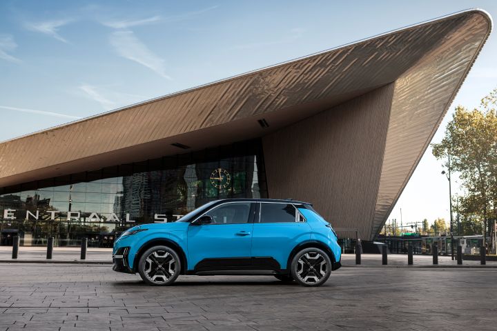 Seitenansicht eines blauen Nissan Micra, der vor dem Hauptbahnhof von Rotterdam steht, dessen reflektierende Dacharchitektur spitz zuläuft