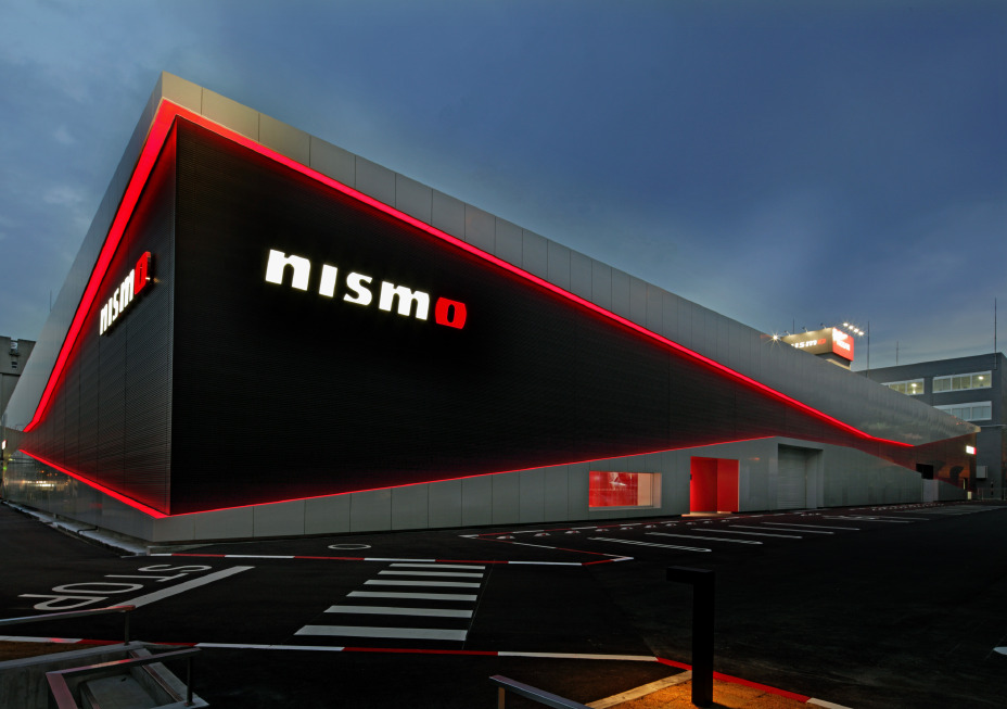 Ein NISMO-Showroom mit schwarz-silbernen Außenwänden vor einem Abendhimmel. Rote LED-Leuchten umrahmen das Gebäude, und an der Vorderseite prangt das „nismo”-Logo in Weiß und Rot. Im Vordergrund befindet sich ein schwarzer Asphaltparkplatz mit weißen Markierungen.