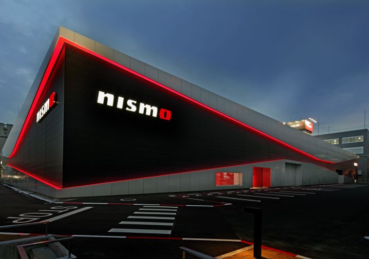 Ein NISMO-Showroom mit schwarz-silbernen Außenwänden vor einem Abendhimmel. Rote LED-Leuchten umrahmen das Gebäude, und an der Vorderseite prangt das „nismo”-Logo in Weiß und Rot. Im Vordergrund befindet sich ein schwarzer Asphaltparkplatz mit weißen Markierungen.
