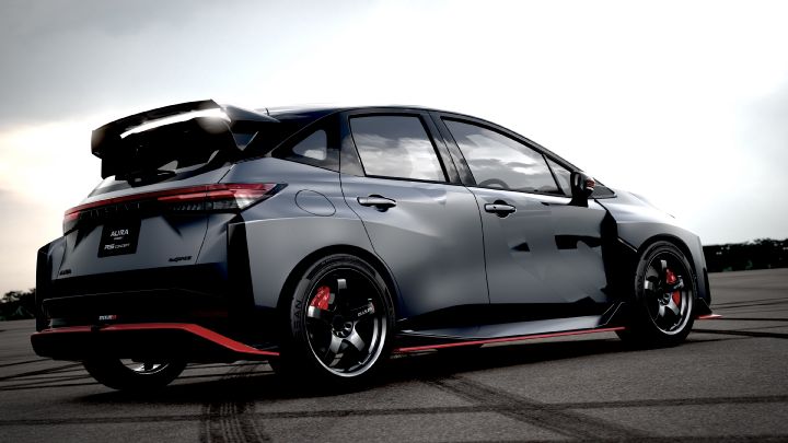 A silver and black two-tone sports car, the Aura Nismo RS Concept, parked on an outdoor skid pad in a rear-right angled view. The vehicle features a large rear wing, with a black plate displaying white text reading 'AURA NISMO RS CONCEPT' at the center of the rear, along with a red accent line and the NISMO logo on the lower bumper. The wheels are black with visible red brake calipers, and the side displays a light gray 'RS.' The background shows a cloudy sky and distant trees, with tire marks visible on the asphalt surface.
