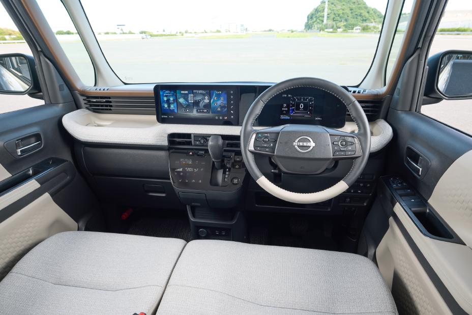 The driver's seat of the new Nissan Roox kei car, showing the steering wheel, digital display, and center console. The outside scenery is also visible through the windows.
