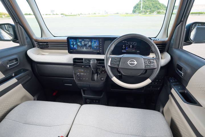 The driver's seat of the new Nissan Roox kei car, showing the steering wheel, digital display, and center console. The outside scenery is also visible through the windows.