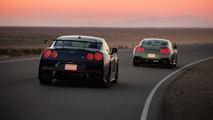 Two Nissan GT‑R sports cars, one black and one silver, drive on a deserted road at sunset. The road curves to the right through a barren landscape with distant mountains under a pink-orange sky.