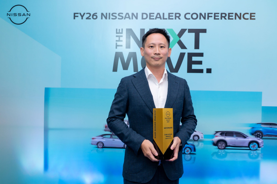 An individual in a business suit stands on a stage holding a yellow award trophy at the FY26 Nissan Dealer Conference, with “The Next Move” displayed on the screen and car images in the background.