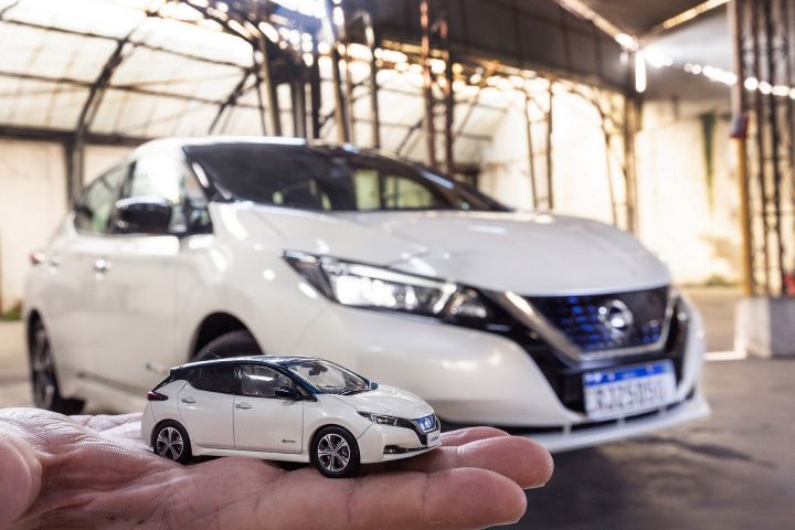 Miniatura de um Nissan Leaf em uma mão com um Nissan Leaf de verdade ao fundo
