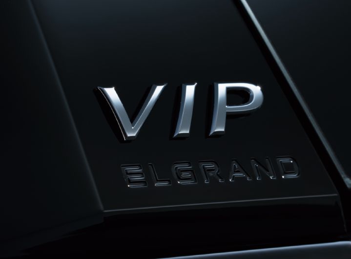 「VIP」および「ELGRAND」のエンブレムをクローズアップで撮影した画像。画面中央には立体的なシルバーの「VIP」エンブレムが大きく配置され、その下に黒い「ELGRAND」の文字が刻まれている。エンブレムは光沢のあるブラックのボディパネル上に取り付けられており、周囲の光を反射して金属的な質感が際立っている。
ボディ表面にはシャープなプレスラインが斜め方向に走り、ブラック塗装ならではの深みと高級感が表現されている。車両の一部を極めて近距離から撮影した構図のため、車両全体の向きは確認できないが、リヤゲートまたはリアピラー付近の加飾エリアを捉えたディテール写真となっている。