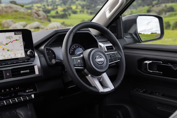 Angled driver-side interior view of a 2026 Nissan Navara ST-X, showing the steering wheel, analogue gauges, part of the 9-inch Touch infotainment screen with navigation displayed, and the climate-control panel below.