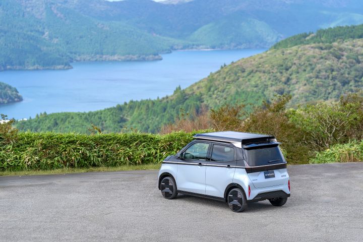 Against a backdrop of a lake and mountains, a light blue Nissan SAKURA is parked by the roadside, facing slightly left rear. The solar roof is closed and rests on top, with green shrubs nearby and a blue lake and mountains in the distance.