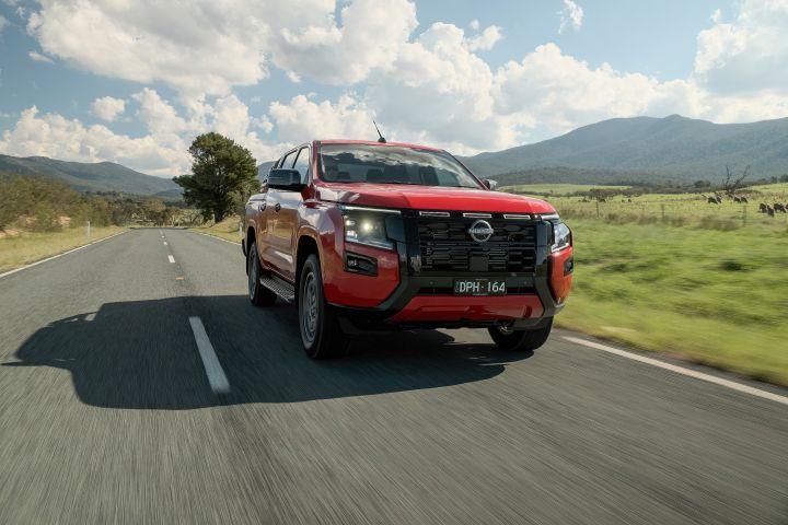 Front three‑quarter view of a 2026 Nissan Navara ST‑X dual cab painted in Outback Red, travelling on a sealed country road lined with green grassy vegetation and distant mountains.