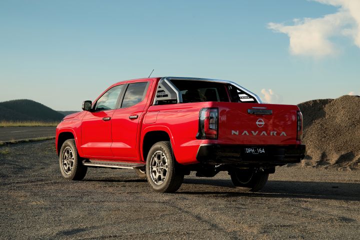 Rear three-quarter view of a 2026 Nissan Navara ST-X dual cab finished in Outback Red, parked on gravel with the sports bar, tailgate, rear lamps, and side steps visible against a mountain backdrop.