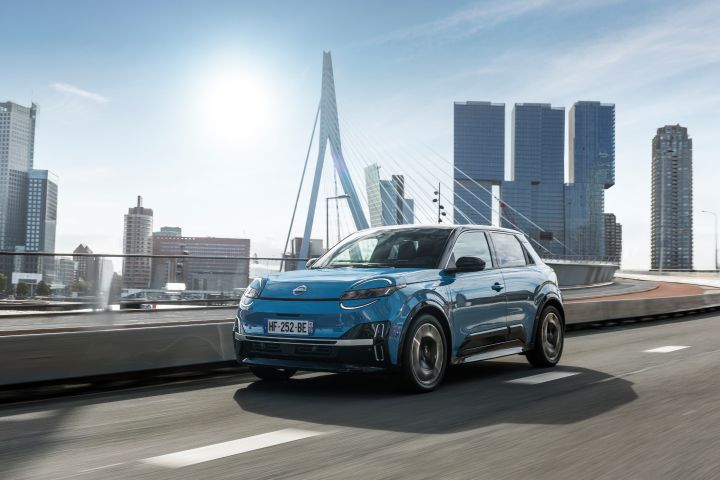A blue Nissan Micra, seen from the front ¾ perspective, drives across a bridge with a city scape in the background