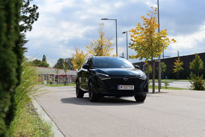 Blick von schräg unten auf die Front eines dunkelgrünen Nissan Qashqai e‑Power, der über einen baumbestandenen Parkplatz fährt